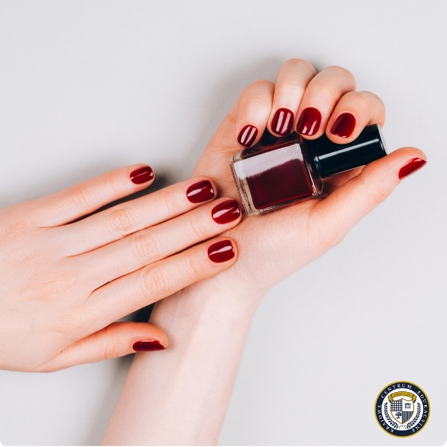 Dłonie z paznokciami pomalowanymi na ciemnoczerwony kolor w stylu manicure klasyczny. Osoba trzyma buteleczkę z lakierem do paznokci w tym samym odcieniu.