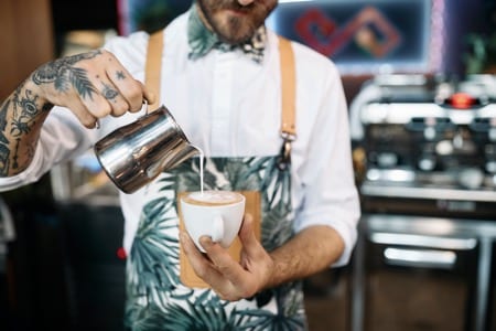 Barista z wytatuowanymi dłońmi nalewający spienione mleko do kawy podczas kursu baristy. Ma na sobie białą koszulę w tropikalne wzory i pomarańczowe szelki, co podkreśla profesjonalny, ale swobodny charakter zawodu