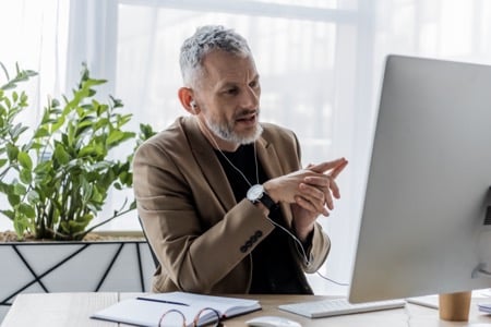 Doświadczony mentor w beżowej marynarce podczas sesji online w ramach kursu coachingu, gestykulujący podczas rozmowy przed monitorem komputera. Na biurku widoczny notatnik i kawa, a w tle rośliny dodające profesjonalnemu wnętrzu przyjaznego charakteru