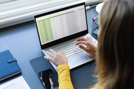 Osoba w żółtym swetrze pracuje na laptopie podczas kursu Excel makra, na ekranie widoczny jest arkusz z kolumnami danych. Na biurku znajduje się niebieski notatnik, a uczestniczka skupiona jest na programowaniu w VBA