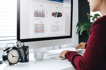 Osoba w bordowym swetrze pracuje przy komputerze podczas kursu Excel podstawowy, analizując wykresy i diagramy wyświetlone na monitorze. Na białym biurku znajduje się również budzik, kubek oraz pojemnik na przybory biurowe