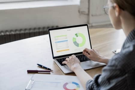 Uczestniczka kursu Excel zaawansowany pracuje na laptopie, analizując złożone wykresy kołowe i diagramy. Na ekranie widoczne są zaawansowane wizualizacje danych, a na biurku leżą dodatkowe materiały szkoleniowe