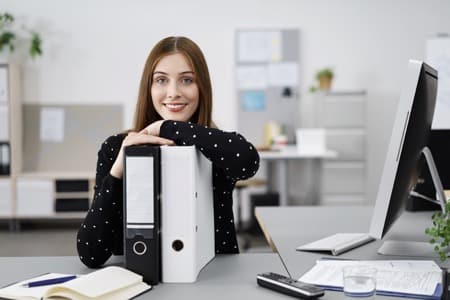 Uśmiechnięta uczestniczka kursu księgowości online pozuje przy biurku z segregatorami dokumentów, komputerem i materiałami księgowymi w nowoczesnym, jasnym biurze, demonstrując profesjonalne podejście do nauki zawodu
