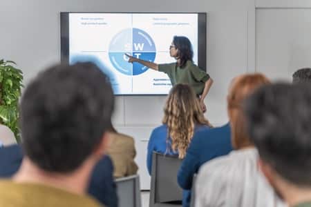 Prowadząca kurs Power Point wskazuje na wyświetlony na dużym ekranie diagram kołowy. Grupa uczestników siedzi na krzesłach, słuchając prezentacji w nowoczesnej sali szkoleniowej wyposażonej w interaktywny wyświetlacz