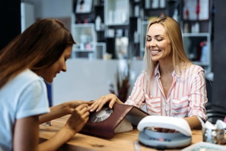Profesjonalistka prowadzi kurs stylizacji paznokci, uśmiechając się podczas praktycznego pokazu technik manicure swojej kursantce w nowoczesnym salonie kosmetycznym wyposażonym w specjalistyczny sprzęt