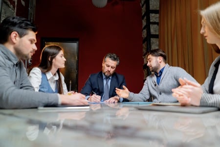 Grupa biznesowa podczas szkolenia z negocjacji, gdzie uczestnicy ćwiczą rzeczywiste scenariusze przy stole konferencyjnym. W centrum mężczyzna w granatowej marynarce prowadzi dyskusję, demonstrując praktyczne zastosowanie technik negocjacyjnych
