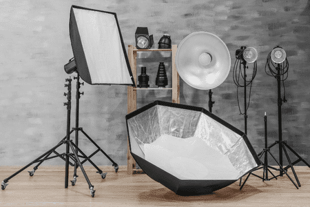 Profesjonalne studio fotograficzne wyposażone w sprzęt niezbędny podczas kursu fotografii - lampy błyskowe, softboxy, modyfikatory światła i akcesoria na drewnianej półce. W tle widoczna szara ściana, a na podłodze parkiet