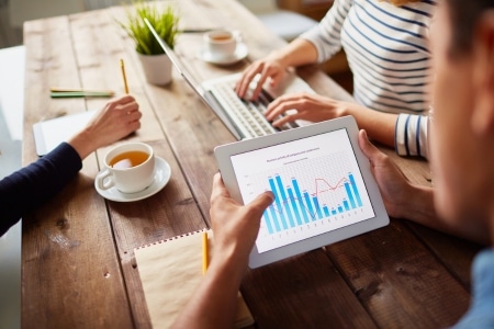 Osoba uczestniczy w kursie marketingu internetowego online, analizując na tablecie wykresy wydajności kampanii. Na drewnianym stole znajdują się filiżanki z herbatą, notatnik i przybory do pisania, podczas gdy inny uczestnik pracuje na komputerze