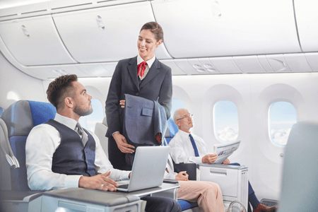 Kurs na stewardessę - szkolenie personelu pokładowego w symulowanej kabinie samolotu. Osoba w służbowym uniformie z czerwonym krawatem demonstruje obsługę pasażerów dwóm kursantom w kamizelkach, z których jeden pracuje na laptopie, a drugi czyta dokumenty. Widoczne są fotele lotnicze, okna samolotu i schowki bagażowe, oddając realistyczne warunki pracy w przestrzeni powietrznej