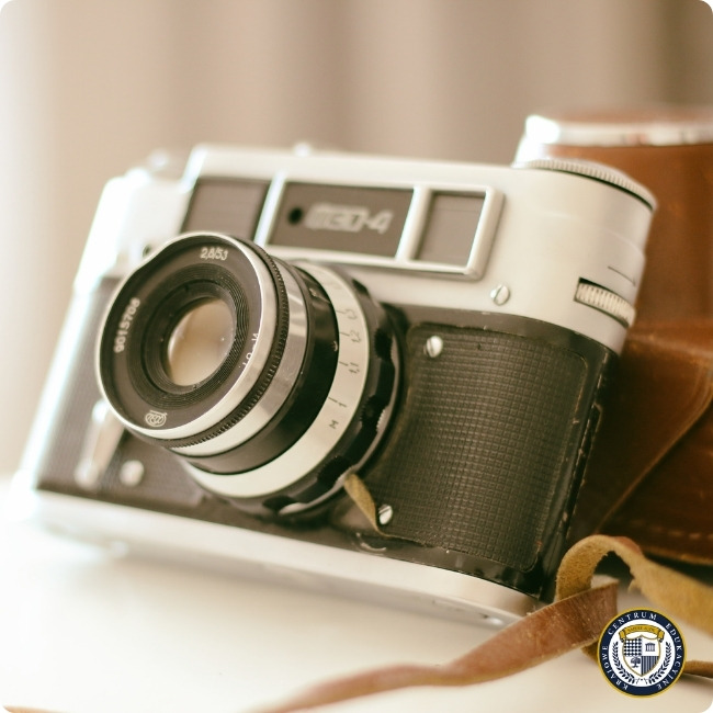 Tradycyjna fotografia: zbliżenie na klasyczny aparat analogowy w biało-czarnej obudowie z widocznym obiektywem i fragmentem skórzanego etui. Zdjęcie utrzymane w ciepłej, nostalgicznej tonacji z delikatnie rozmytym tłem