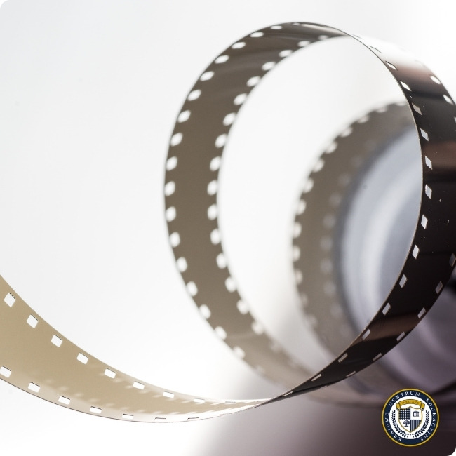 Artystyczne ujęcie rozwiniętej taśmy filmowej 35mm z charakterystycznymi perforacjami na krawędziach. Taśma tworzy elegancką, zakrzywioną linię na białym tle, podkreślając analogowy charakter fotografii