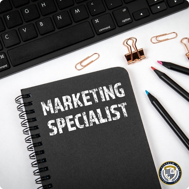 Specjalista ds. marketingu zarobki: Czarny notes z napisem 'MARKETING SPECIALIST' na biurku obok klawiatury komputerowej. Na białym blacie rozłożone różowe spinacze biurowe i kolorowe markery