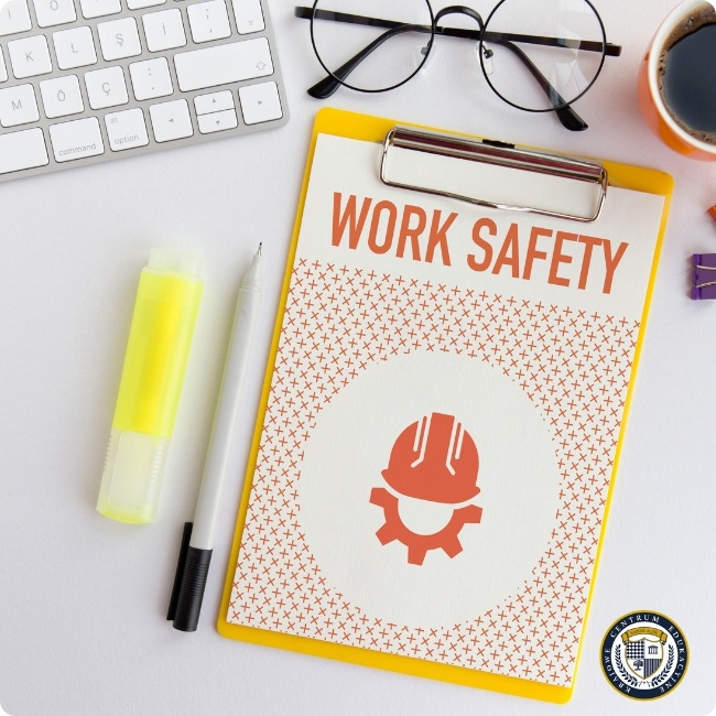 Tekst alternatywny (alt text):"Widok biurka z góry z dokumentem zatytułowanym 'WORK SAFETY' na żółtym clipboardzie. Na dokumencie widnieje ikona kasku ochronnego w pomarańczowym kolorze. Na biurku znajdują się również: biała klawiatura komputerowa, okrągłe okulary, zakreślacz, długopis, filiżanka kawy oraz fioletowy spinacz. Zdjęcie przedstawia elementy związane z bezpieczeństwem pracy w przestrzeni biurowej
