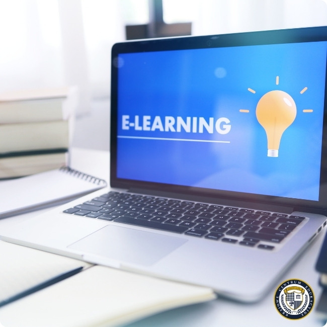 Ekran laptopa wyświetlający napis 'E-LEARNING' na niebieskim tle z ikoną żółtej żarówki. Obok laptopa widoczny notes i inne materiały do nauki na białym biurku