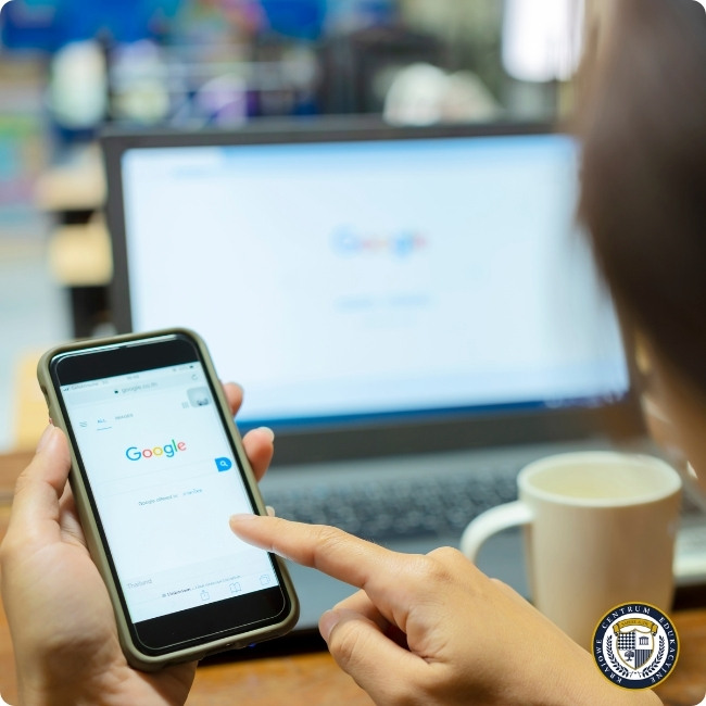 Osoba pracująca jednocześnie na laptopie i smartfonie z otwartą wyszukiwarką Google, obok stoi kubek z napojem. W prawym dolnym rogu widoczne logo KCE. Zdjęcie pokazuje pracę w nowoczesnym środowisku biurowym