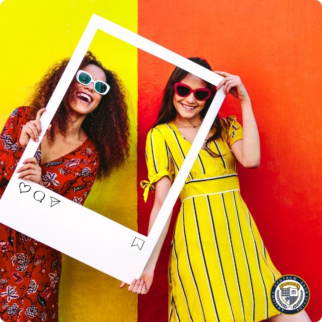  Dwie uśmiechnięte osoby pozujące z białą ramką w stylu Instagram, na kolorowym tle żółto-czerwonym, ubrane w wzorzyste sukienki i okulary przeciwsłoneczne